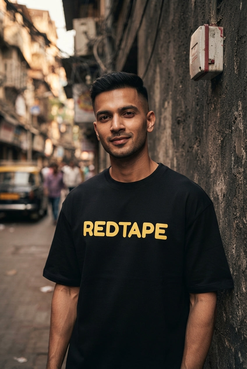 A young Indian man wearing a black oversized REDTAPE printed cotton t-shirt standing against a charcoal concrete urban wall.